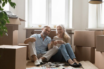 Mature couple sit on floor near cardboard boxes with stuff on relocation day, preparing for repairs...