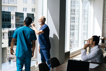 Rear view of team multi-ethnic doctors discussing together history disease of patient using MRI scan during medical conference standing near window. Concept of modern medicine and health care.