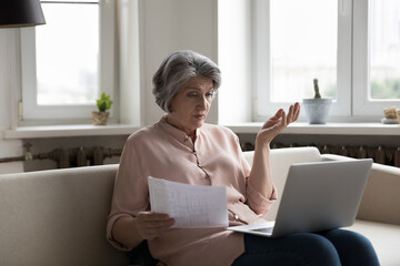 Older woman sit on sofa put wireless computer on laps, check bills, experiences problems with...