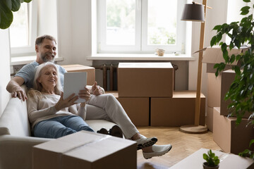 Mature couple using digital tablet buying goods for new home relaxing on cozy couch in living room with packed cardboard boxes with stuff on relocation day. Do easy comfort remote e-shopping concept
