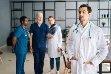 Portrait of thoughtful young male doctor wearing white uniform standing in medical meeting office, looking away. Multi-ethnic team of physician working in background, discussing xray scan of patient.