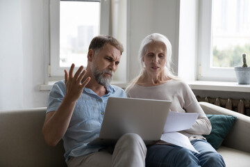 Elderly couple sit on sofa at home with laptop sorting out papers, reviewing expenses, feel angry due to utility bills going up. Financial crisis, bank debt notice, bankruptcy, lack of money concept