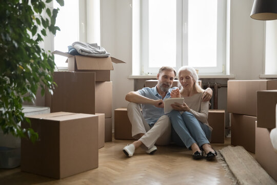 Mature spouses shoppers sit on floor near heap of cardboard boxes on relocation day use tablet buy furniture for new home through online web store. E-shopping, retail application, modern tech concept