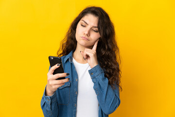Teenager Russian girl isolated on yellow background using mobile phone and thinking