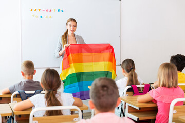 Young progressive female teacher discussing with preteen children about LGBT social movements in classroom, holding rainbow flag - obrazy, fototapety, plakaty