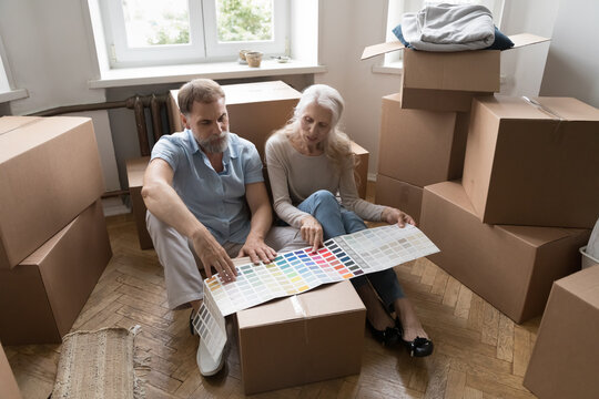 Elderly couple review paint swatches palette sheet, choose wall colour prepare house for repairs works sit on floor in living room near boxes with personal stuff on relocation day. Renovation concept