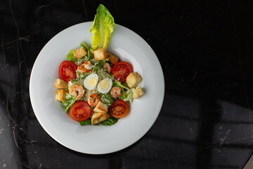 Caesar salad with grilled shrimps, tomato, boiled egg, lettuce, croutons and green sauce. Top view close-up on a brown round plate with fresh vegetables and cutlery by side on a dark background.
