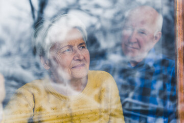 Shot from behind the window glass. Concerned gray-haired elderly woman looking out of the window,...