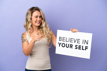 Young Brazilian woman isolated on purple background holding a placard with text Believe In Your Self and pointing to the front