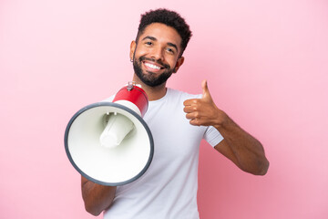Young Brazilian man isolated on pink background holding a megaphone with thumb up
