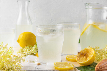 Fresh iced lemonade with lemons and elderberry flowers in a jar and glasses on white tile table.