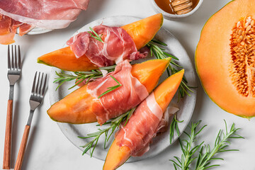 Prosciutto ham with melon cantaloupe slices, honey and rosemary in a plate with forks on white...