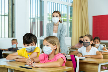 Fototapeta premium Focused preteen pupils in protective face masks studying in classroom with female teacher. Necessary precautions in coronavirus pandemic