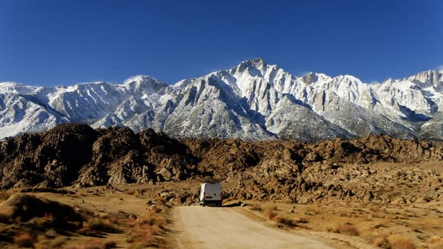 Aerial view of a caravan driving on a dirt road with snow topped mountain background, in USA - circling, drone shot