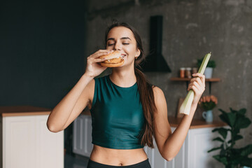 Sporty woman choosing between vegetable and burger while standing on light kitchen. Woman eating...