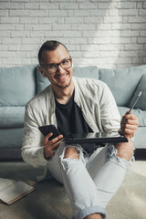 Smiling man in eyeglasses working remotely with digital tablet and mobile phone while sitting on floor at home. Business communication. Mobile communication