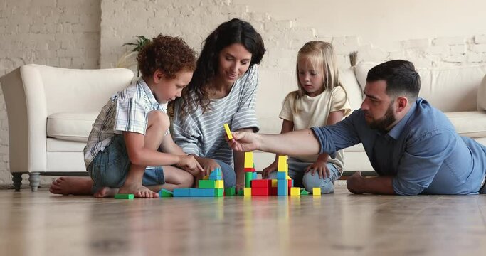 Happy family foster Latin mom dad two small adopted children son daughter relax on warm floor enjoy playing constructor set. Couple of parents with kids create fantasy town from wooden colorful blocks