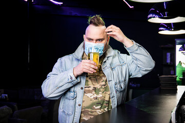 punk man with a mohawk wearing a protective mask on his face drinks beer from a glass mug in the...