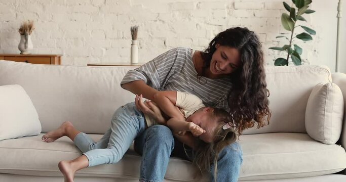 Affectionate millennial single mother female babysitter play with active little daughter tickle laughing girl relax having fun together with child. Happy mom mess with joyful kid on large cozy couch