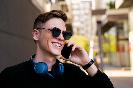 Handsome Young Guy Talking On Smart Phone With Teeth Smile, Headphones Around His Neck 