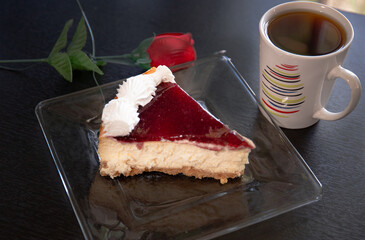A slice of cheesecake in a glass plate with a coffe cup and a rose
