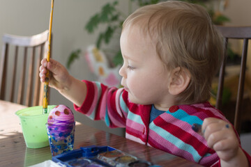 Toddler in a pink striped sweater paints an Easter egg with watercolor paint; toddler holds a large paint brush in her hand