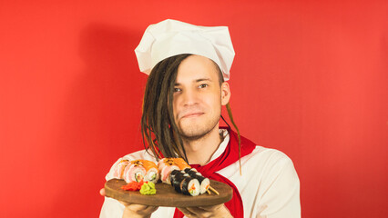 Young man dressed as chef holding wooden board with sushi rolls. Male cook with appetizing sushi rolls on red background. Concept of fast food and takeaway.