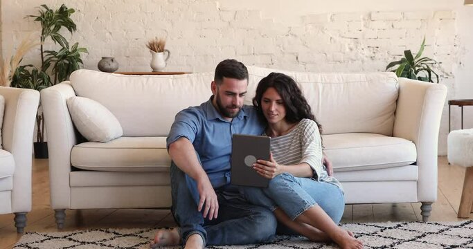 Romantic young couple spouses or lovers relax on carpet at living room use modern electronic tablet pc. Happy millennial man woman in love sit on warm floor cuddle do shopping networking online on pad