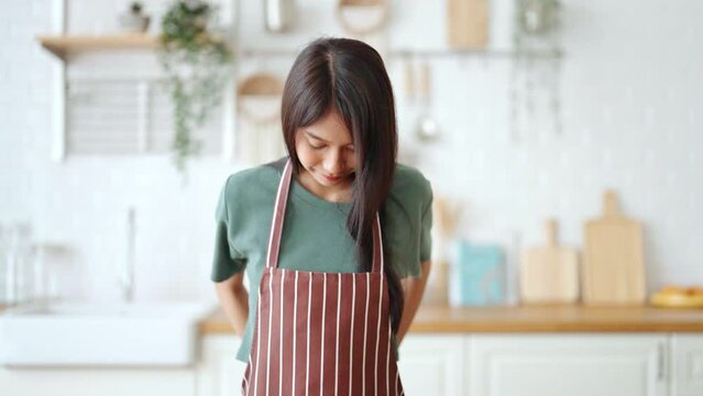 Happy young asian woman wearing apron and standing in kitchen room. Beautiful female smiling and looking at camera feeling confident