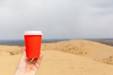 A red cup of coffee or a hot drink, covered with a lid in your hand against the background of a desert or a sandy seashore.