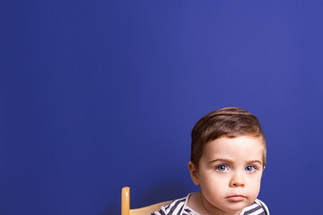 Thoughtful cute toddler boy on chair on blue background.Baby portrait isolated copy space