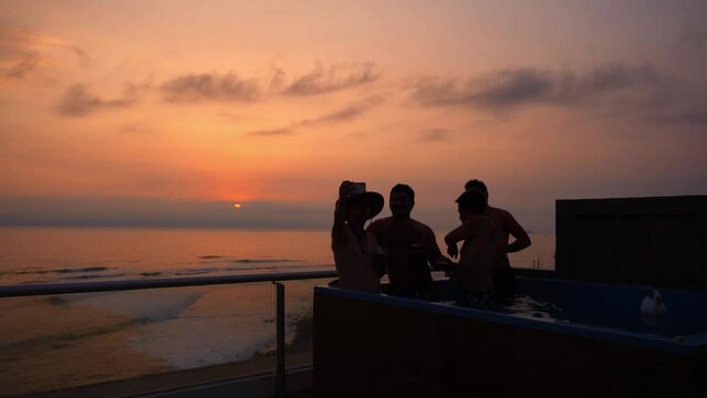 several friends taking a selfie with a cell phone with orange sunset with glasses of beer drink in a swimming pool in a house with a view of the beach in 4k