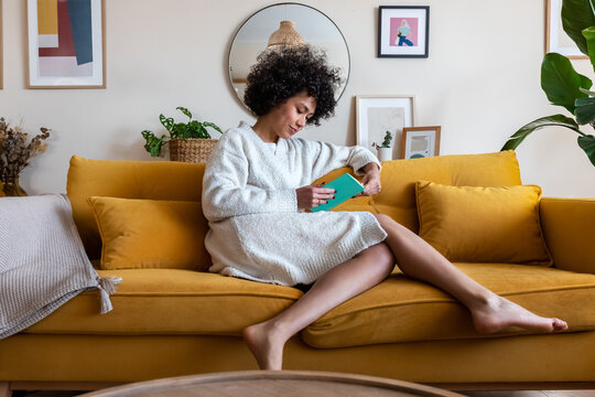 African american woman relaxing at home reading a book sitting on couch.