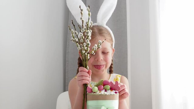 A beautiful, cute, smart girl in an Easter bunny costume smiles and holds a willow bouquet in her hand. symbol of the holiday of light easter - painted eggs, easter cake, willow, easter rabbit.
