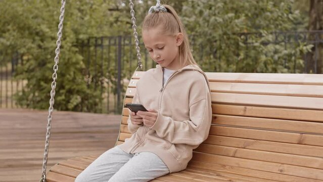 Slowmo of pretty 10 year old Caucasian girl smiling while looking at smartphone screen, sitting on wooden swing bench at playground on sunny day