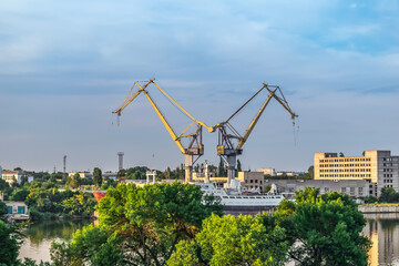 Evening cityscape of Mykolaiv with two port cranes on the horizon, Ukraine. Green trees against the background of the Inhul River with ship and buildings on the shore