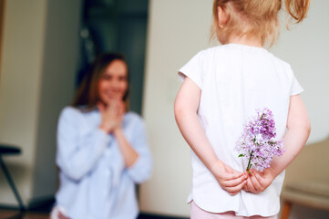 Fototapeta premium Mothers Day. A little preschool girl holds a bouquet of lilac flowers behind her back and wants to give it to her mom