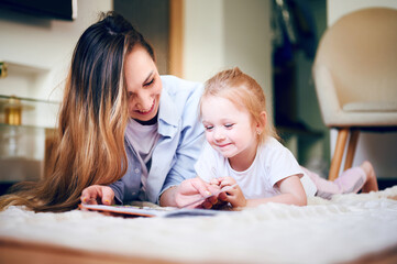 Young mother with her cute little daughter is reading book and looking at the drawings in the book while lying on the floor. The mother teaches the child. The relationship between parents and children