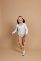 Fototapeta na wymiar Portrait of young beautiful happy smiling girl with long dark hair in white training swimsuit and socks standing on brown background, posing, having fun. Healthy lifestyle, initial gymnastics, sport, 