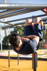 Obraz premium Back view of sportsman working out on sunny day. Man sportive clothes on open air sports ground, doing upside down sit-ups on bars. Sport, health, working out concept