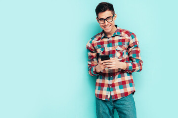 Handsome smiling model.Sexy stylish man dressed in checkered shirt and jeans. Fashion hipster male posing near blue wall in studio. Holding smartphone. Looking at cellphone screen. Using apps