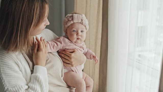 Side view of young brown-haired mother holding cute Biracial baby girl in pink hat at home in afternoon, standing by window at home