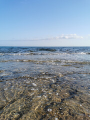 The coast of the Mediterranean Sea, waves, clear water through which the bottom is visible. All this against a blue sky with clouds.