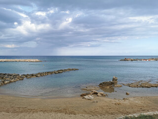 Fototapeta na wymiar Bay of the Mediterranean Sea, sandy shore with a stone ridge and stones against a dramatic sky.
