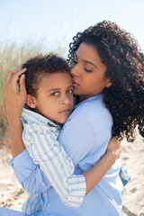 Gentle African American mother and child on beach. Mother and son in casual clothes sitting on blanket, hugging, eyes closed. Family, relaxation, nature concept