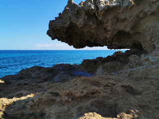 A stone, a rock, a hanging piece of long-frozen lava, below dried salt, similar to ice, against the background of the sea and a blue cloudless sky.
