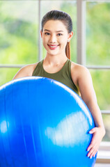 Naklejka premium Millennial Asian young healthy slim strong sexy female sporty model in casual crop top sportswear sitting smiling posing holding hand on exercise ball on yoga mat ready to workout in classroom studio