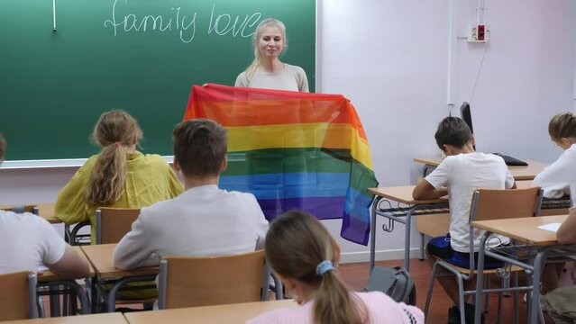 Female teacher holding rainbow-colored LGBT community flag and explaining it meaning for kids during lesson dedicated to family love.