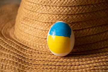 One white chicken egg with a painted flag of Ukraine lies on a straw hat on the table at home,...