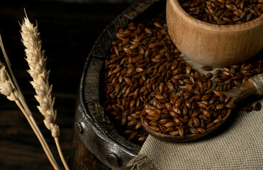 Barley malt for dark beer on a barrel in a bowl with a spoon, dark background. Ingredient for beer.